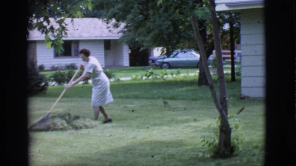 1966 Clarkdale, Arizona home-movie frame of a woman in a light dress raking grass in a yard, with trees, a house, and a parked car behind her.