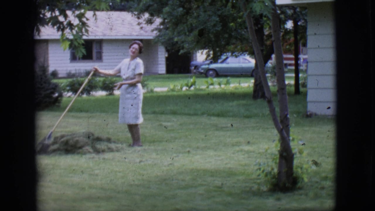 The woman pauses with the rake and looks toward the camera in a 1966 Clarkdale yard, with trees, lawn, house, and parked car visible.