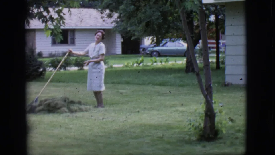 The woman pauses with the rake and looks toward the camera in a 1966 Clarkdale yard, with trees, lawn, house, and parked car visible.