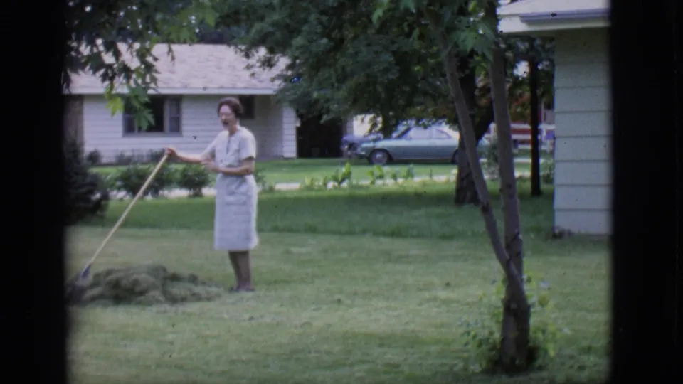 A woman in a light dress stands with a rake in a Clarkdale residential yard, captured in 1966 home-movie footage.