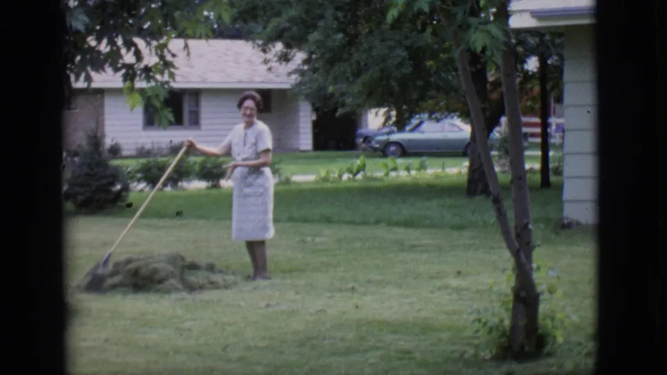 The woman smiles toward the camera while holding a rake in the 1966 Clarkdale yard scene, with lawn, trees, house, and car behind her.