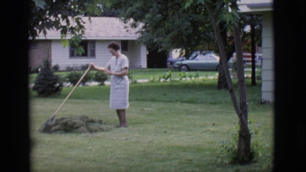Woman in a light dress rakes grass clippings on a front lawn.