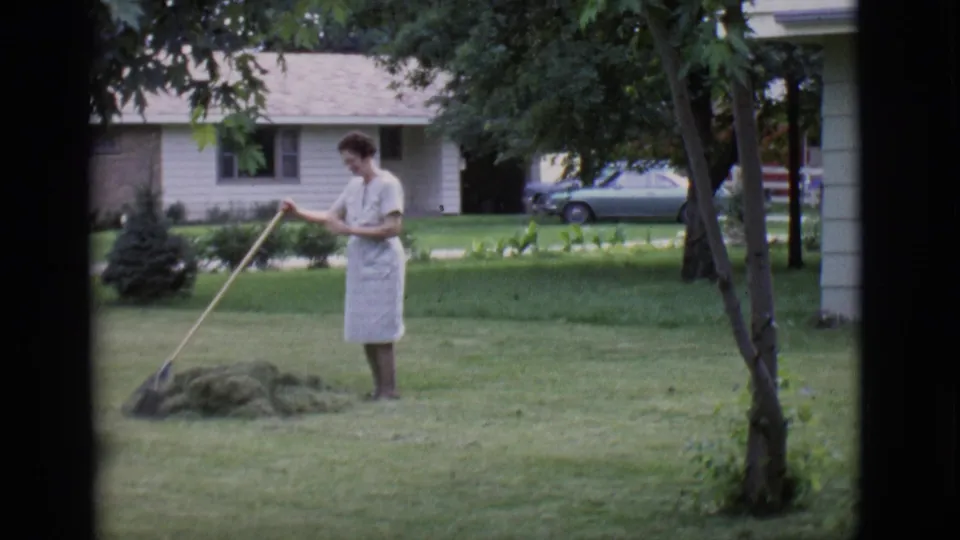 In Clarkdale, Arizona, a woman in a light dress and apron rakes grass clippings on a green residential lawn in 1966.