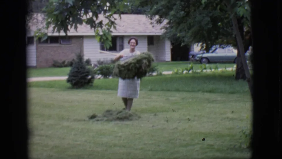 A woman in a light dress reacts toward the camera while holding a large bundle of grass clippings in a Clarkdale yard.