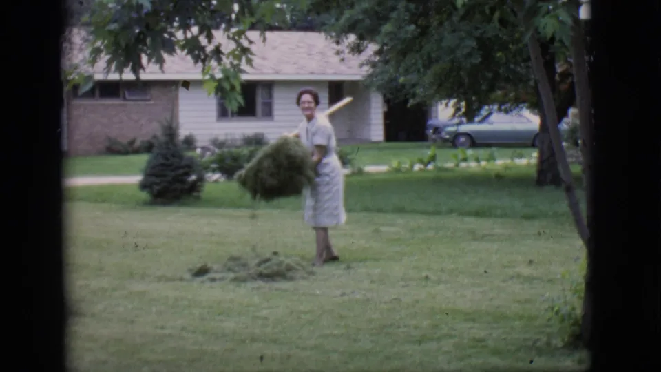 The woman leans over the grass pile with a rake nearby, still looking toward the camera in the 1966 Clarkdale lawn scene.