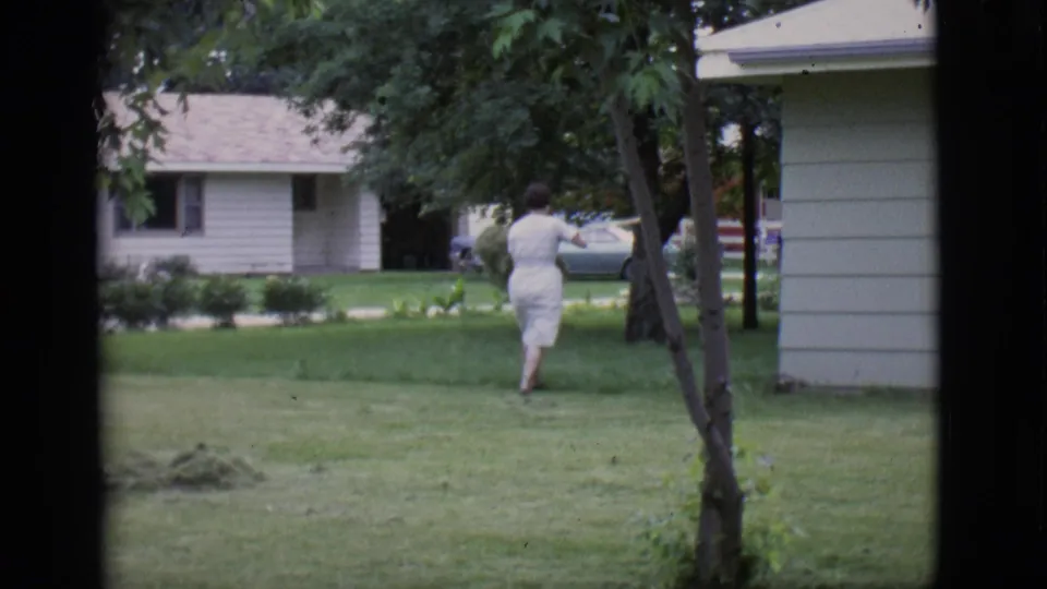 The woman continues across the grassy residential yard, moving toward trees and houses while carrying the clippings.