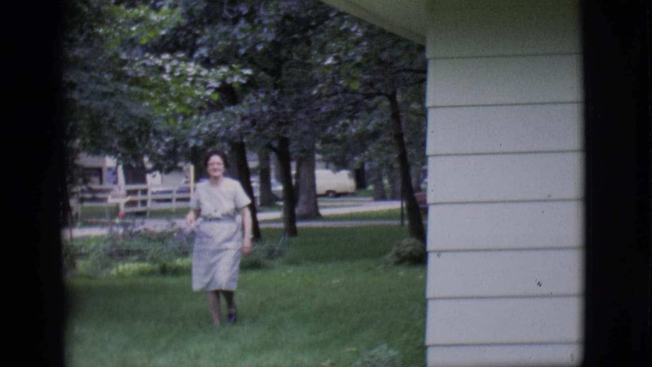 In 1966 Clarkdale, an adult woman in a light dress walks through a residential lawn while holding a rake.