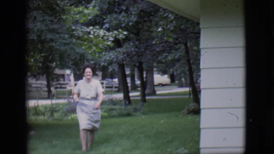 Adult woman with glasses approaches the camera across a Clarkdale lawn, carrying a rake in a 1966 home-movie frame.