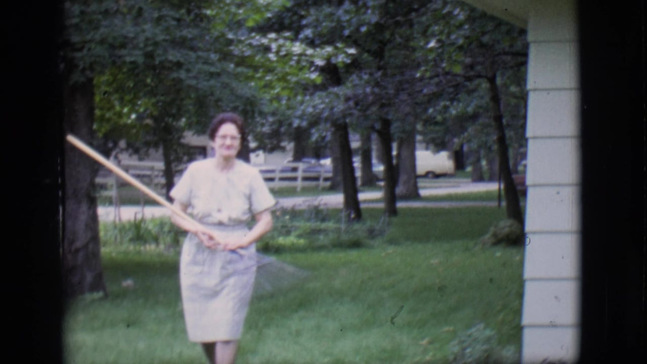 Older woman in a light dress holds a rake while standing on a green lawn.