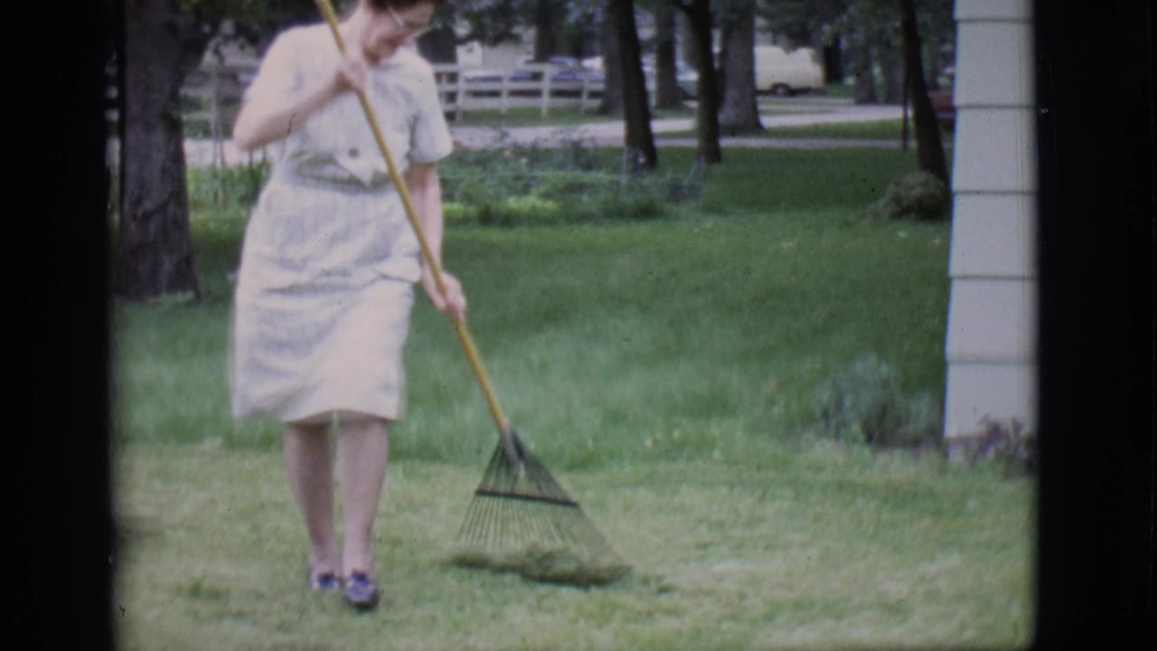 Woman rakes cut grass on a lawn beside a house and trees.