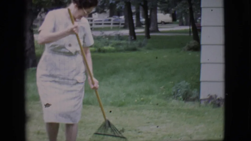 A closer 8mm frame shows the woman looking down as she rakes the backyard lawn, with trees and house siding in the background.