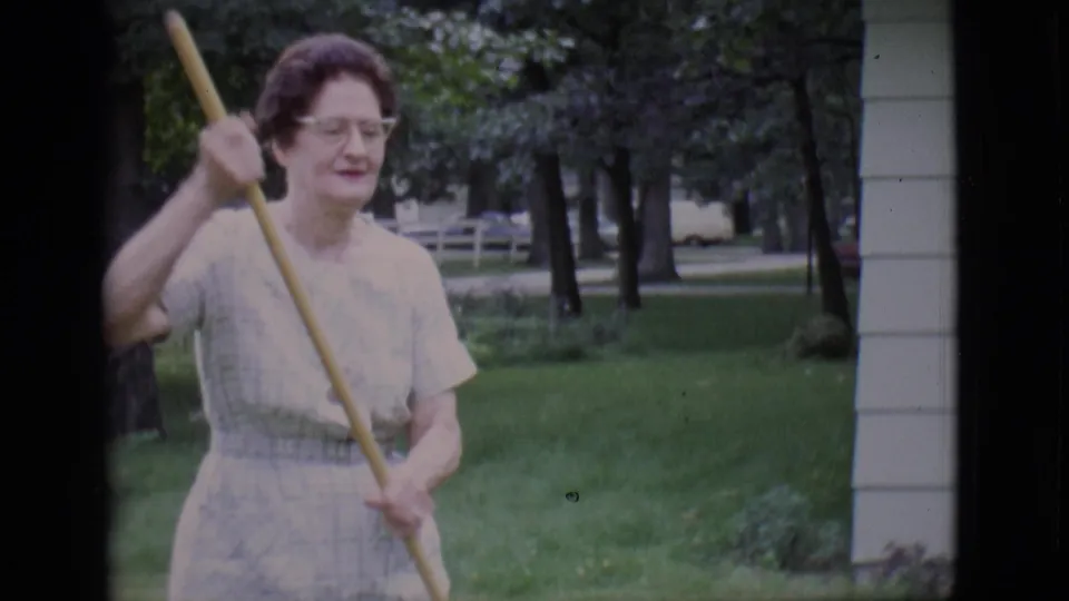 The woman, wearing glasses and a light dress, holds the rake upright in the green backyard during the 1966 Clarkdale home-movie scene.