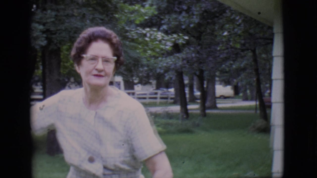 Woman with glasses stands near a porch and lawn with trees behind her.