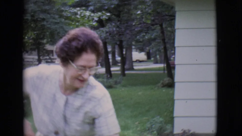The woman looks down while standing near porch siding and a green yard in the 1966 Clarkdale home-movie scene.