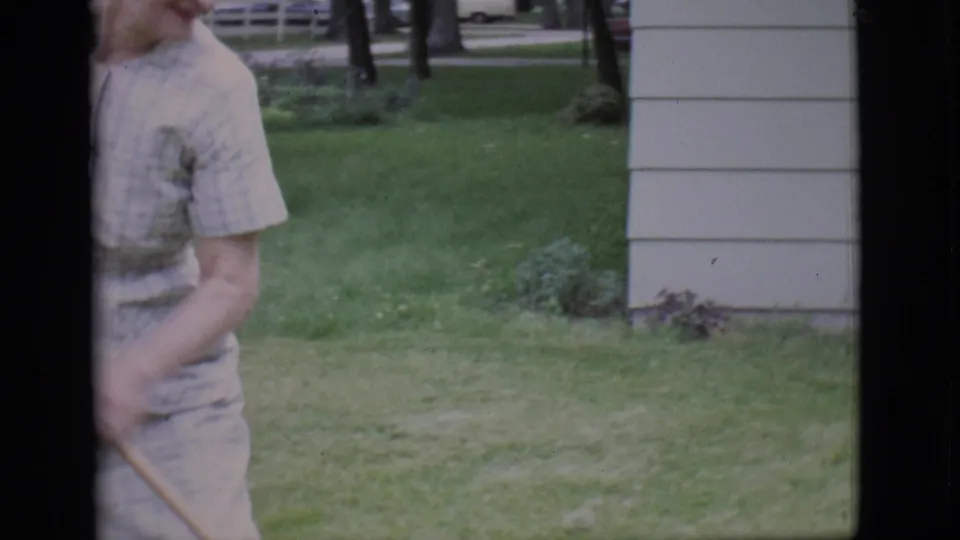 A partial frame shows the woman with a rake beside the lawn and house siding, consistent with the yard-care action described.