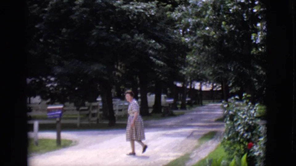 The woman walks toward or past a roadside mailbox on a tree-lined driveway in the Clarkdale, Arizona home-movie footage.