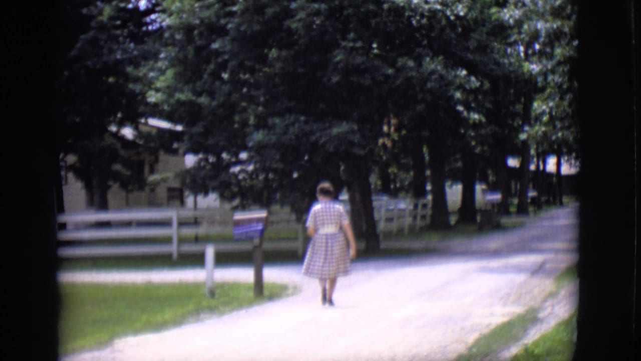 Woman walking away along a curved gravel road beside trees and fencing.