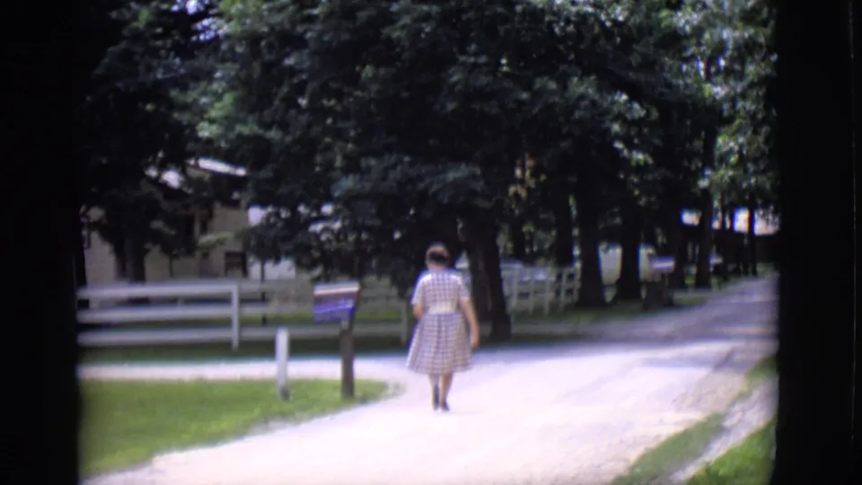 The woman continues away from the camera on a sunlit gravel road in 1966 Clarkdale, with trees and a white fence nearby.