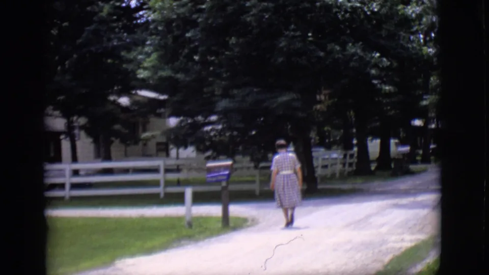 A woman in a mid-century dress walks farther down the Clarkdale gravel road, with a white fence and shaded house behind her.