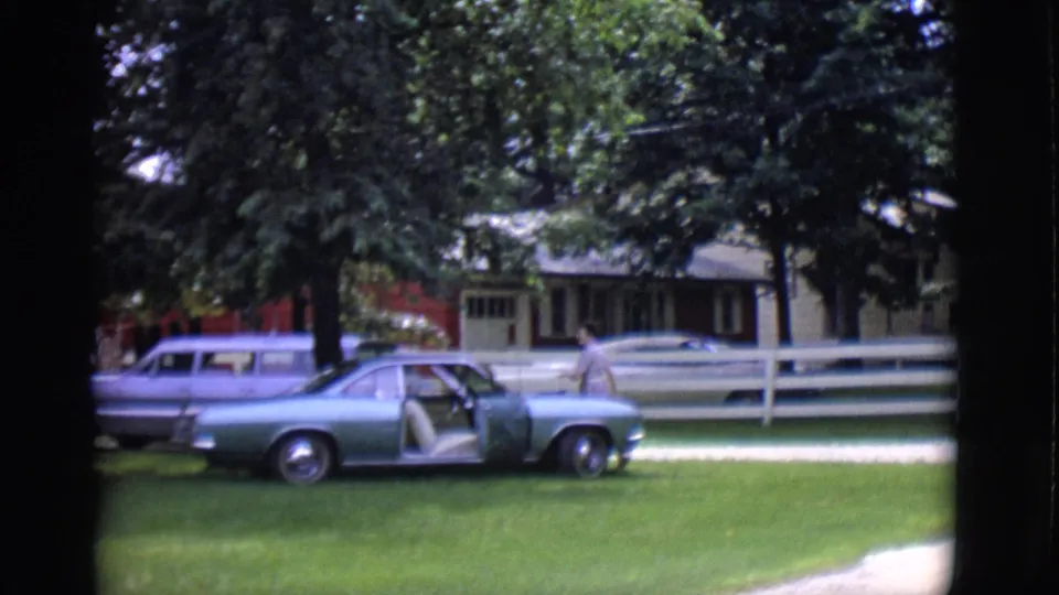 A light blue classic car sits on the lawn with its door open while a station wagon is visible behind it near the house.
