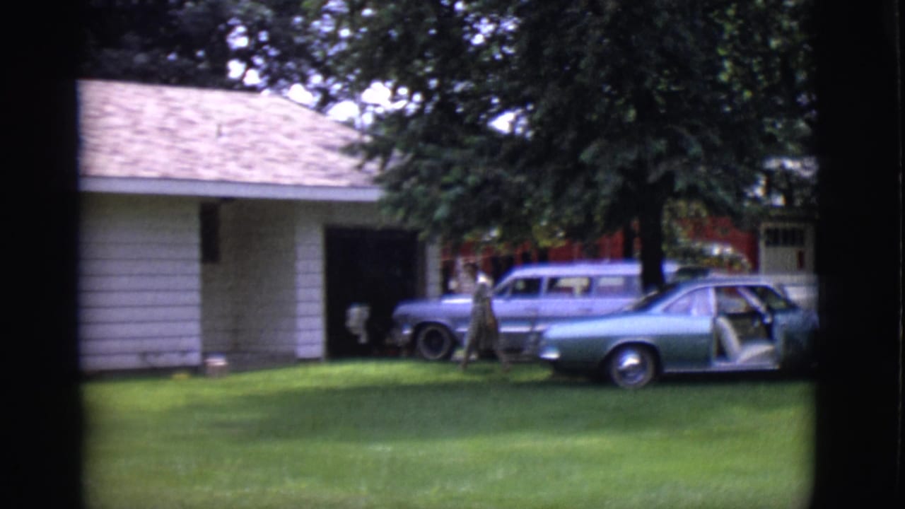 In Clarkdale, Arizona, a woman crosses the lawn near two parked cars outside a white-sided house in this 1966 8mm frame.