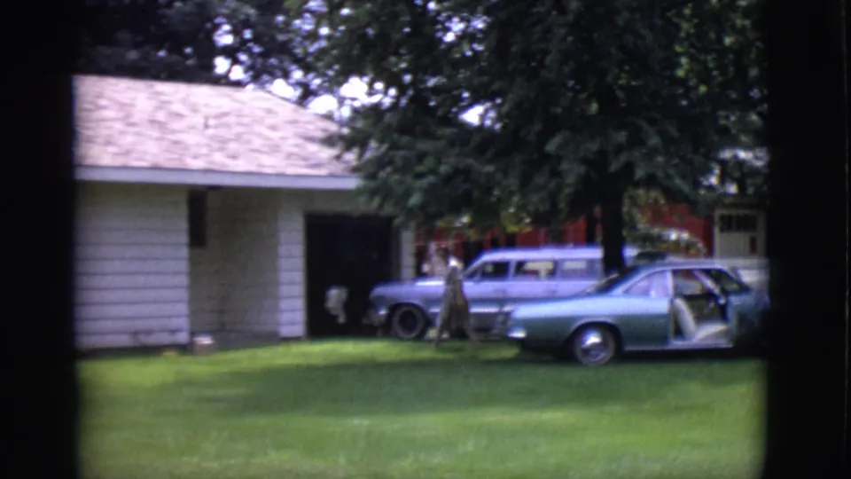 In Clarkdale, Arizona, a woman crosses the lawn near two parked cars outside a white-sided house in this 1966 8mm frame.
