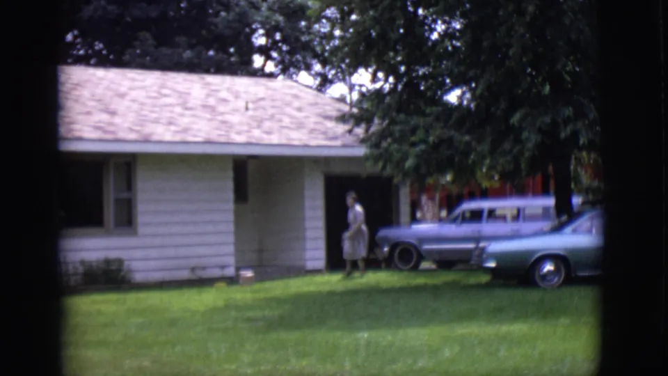 A woman in a light-colored dress moves across the grass toward the white-sided house, with parked classic cars partly visible nearby.