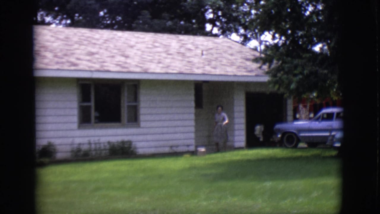 Woman standing near the front entrance of a ranch-style house with a blue station wagon in the driveway.