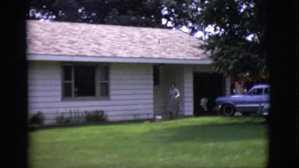 1966 Clarkdale, Arizona home-movie frame showing a woman by a white ranch-style house and a blue station wagon on a green lawn.