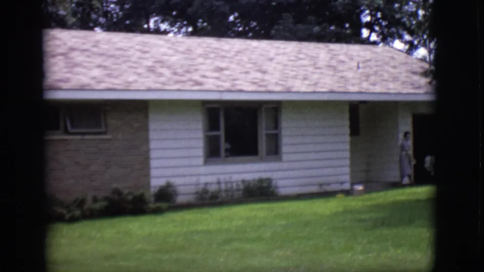 1966 Clarkdale, Arizona home-movie frame showing the house exterior, brick accent wall, lawn, and woman near the front entrance.