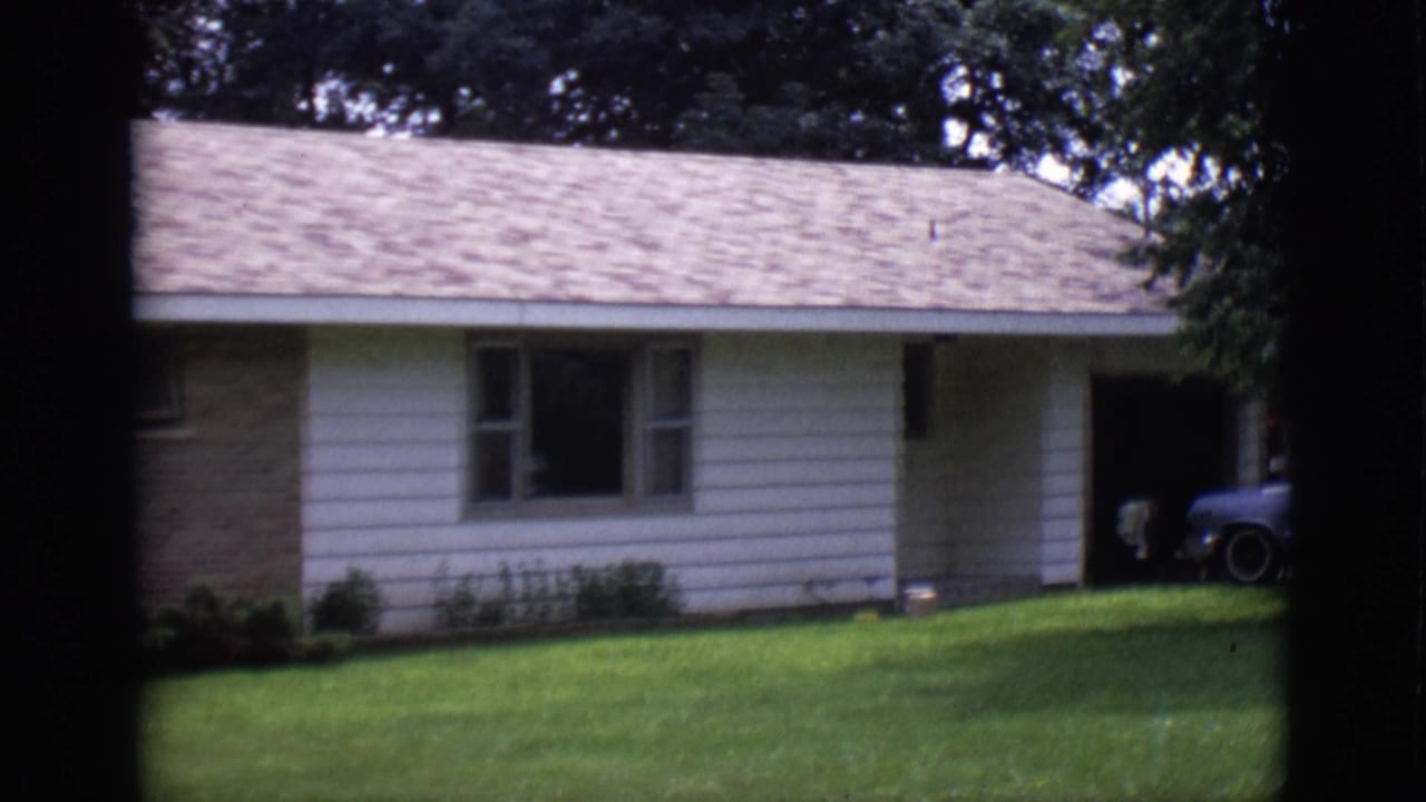 House front with lawn, recessed entry area, trees, and part of a parked car at the driveway edge.