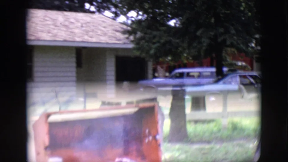 Clarkdale, Arizona 1966 frame with motion blur as the pan passes a tree, front lawn, driveway, and parked station wagons near the house.