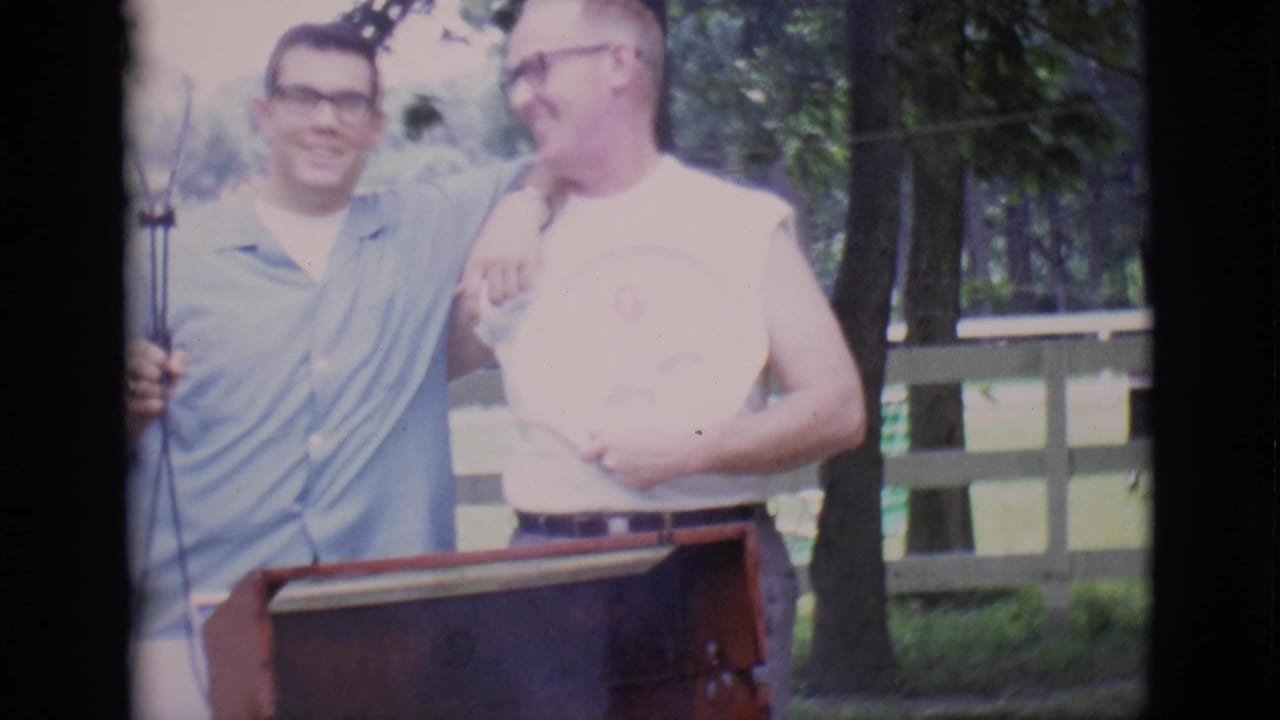 Two men stand behind a red barbecue grill outdoors, one holding a microphone stand.