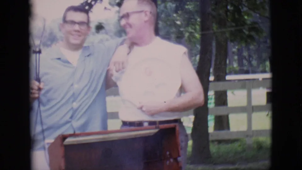 In Clarkdale, Arizona, 1966, two men in casual shirts smile behind a red rotisserie grill in a grassy yard.
