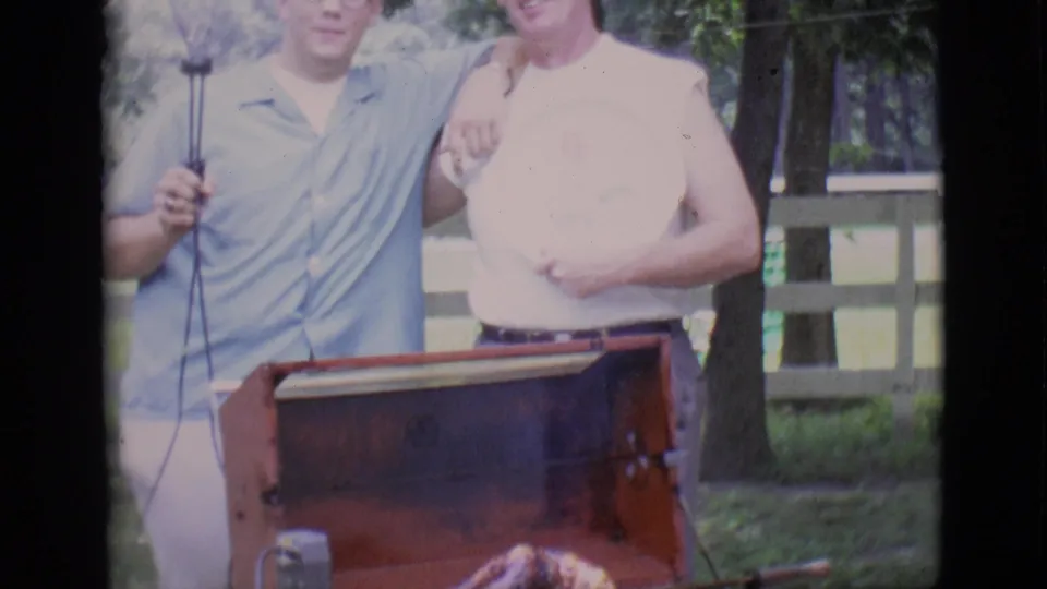 Two men in casual 1960s clothing stand behind a red barbecue grill as a roast turns outdoors in Clarkdale.