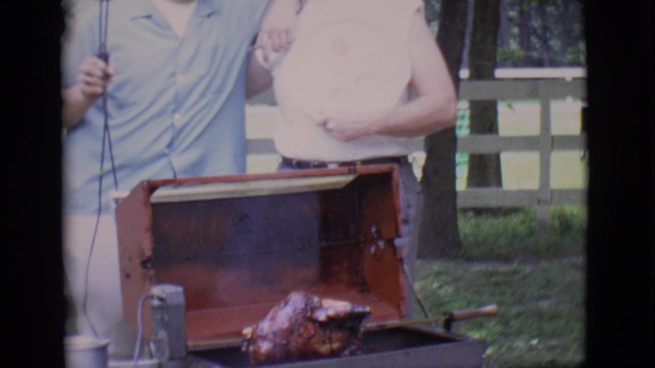 Two men stand beside an open barbecue with a rotisserie chicken in a garden.
