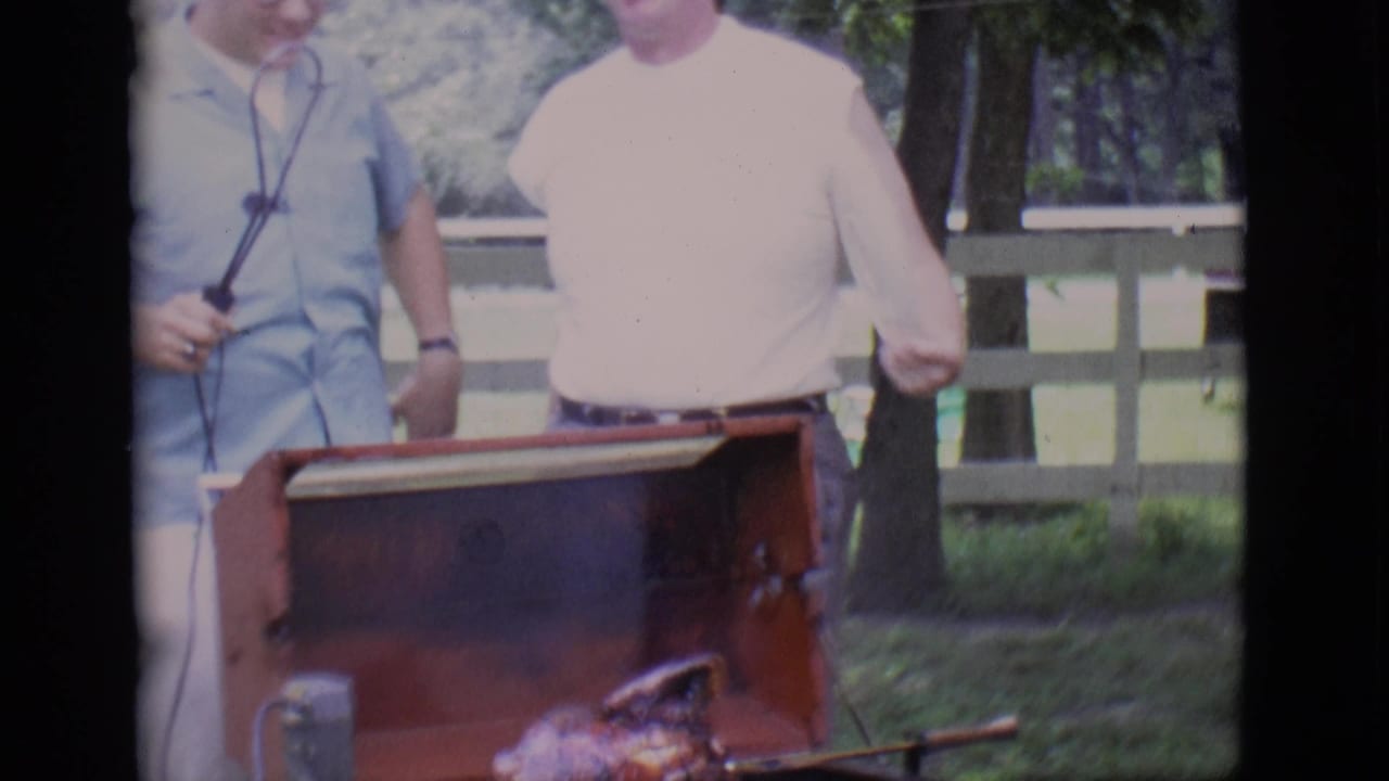 Two men remain beside the open barbecue in the 1966 Clarkdale garden scene, with one holding the spit handle near the cooking chicken.