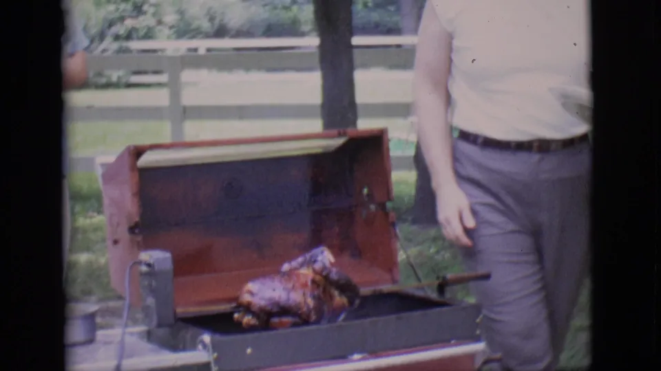 The frame shows the open portable barbecue and rotisserie chicken, with a man partly visible beside the grill in the Clarkdale garden.