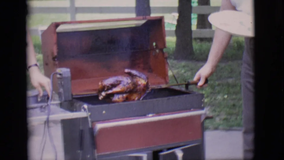 A person turns the rotisserie spit on the open barbecue, keeping the chicken over the grill in the 1966 backyard scene.