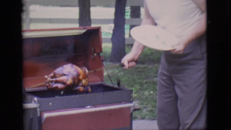The turkey sits on the rotisserie spit inside the open backyard grill while the man stands close with a plate partially visible.