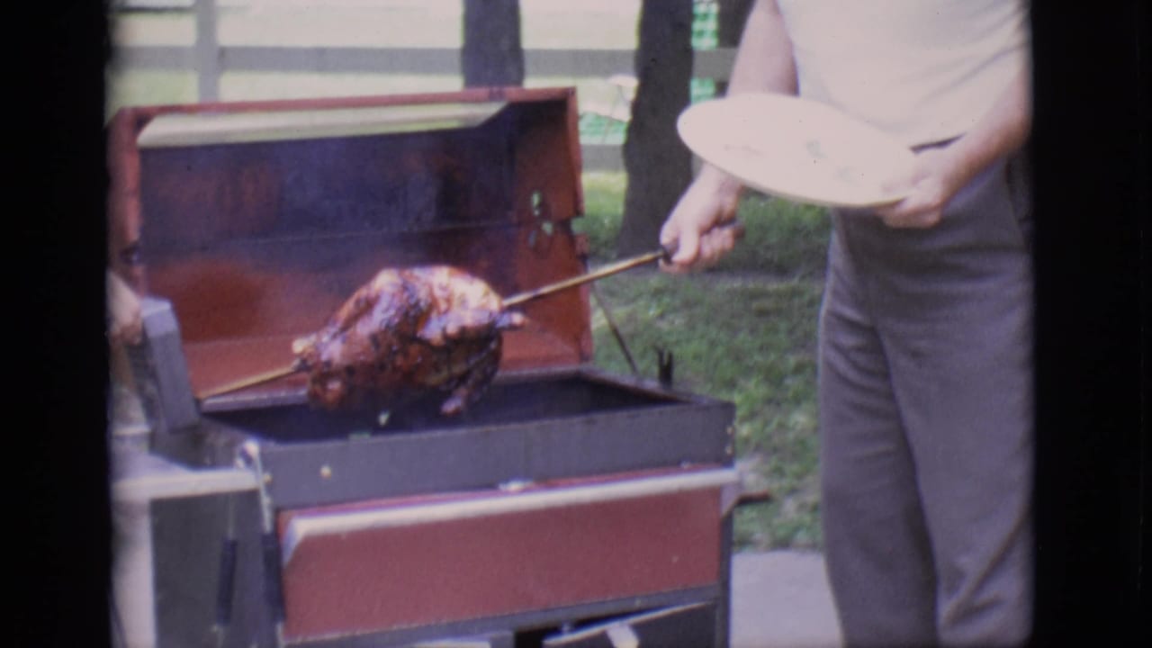 Man uses a metal rod to hold a roasted turkey over an open red grill.