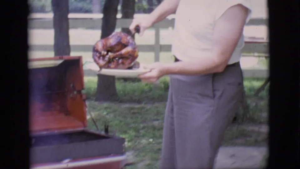 In the backyard scene, the man transfers the roasted turkey from the metal rod onto a white plate near the open red grill.