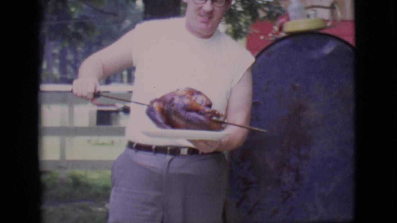 Man in a sleeveless white shirt holds a cooked bird on a plate beside a red grill.