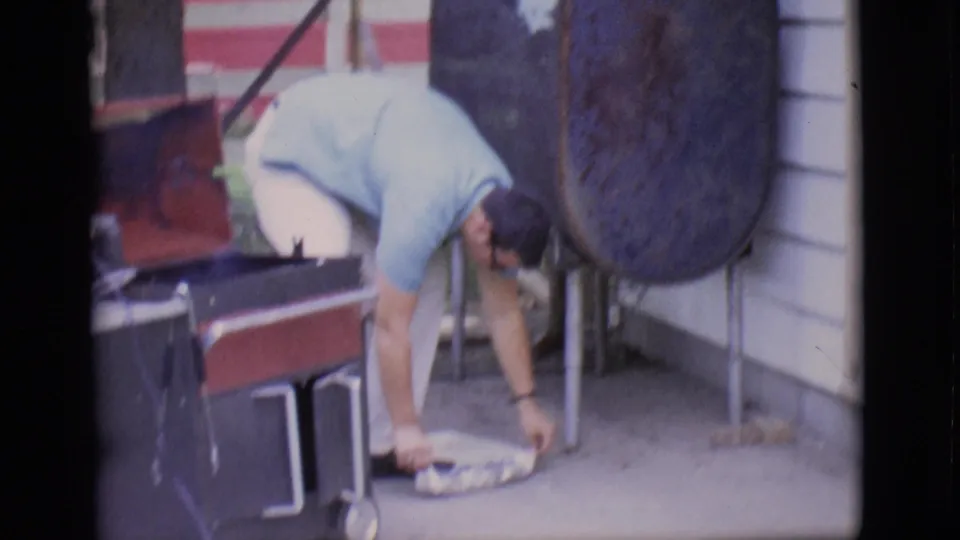 The man bends over a plate on the patio beside the grill setup and house siding in the 1966 Clarkdale footage.