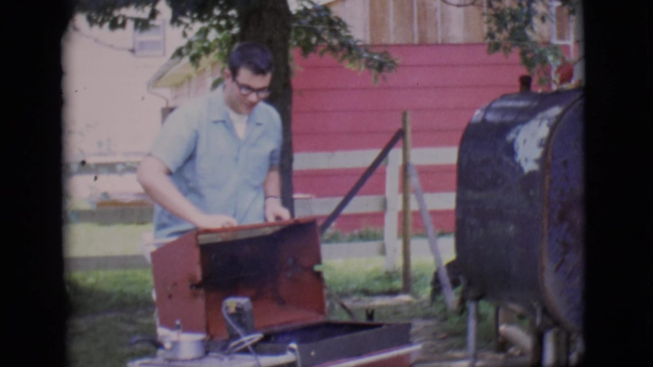 Man in glasses works at an open red grill in a shaded yard.
