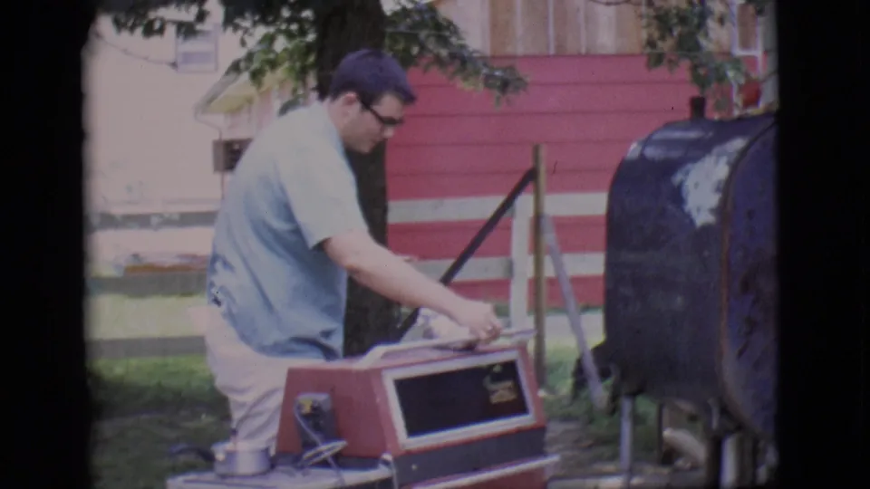 A man in a pale blue shirt reaches across a red backyard grill, with the black smoker and red house siding nearby.