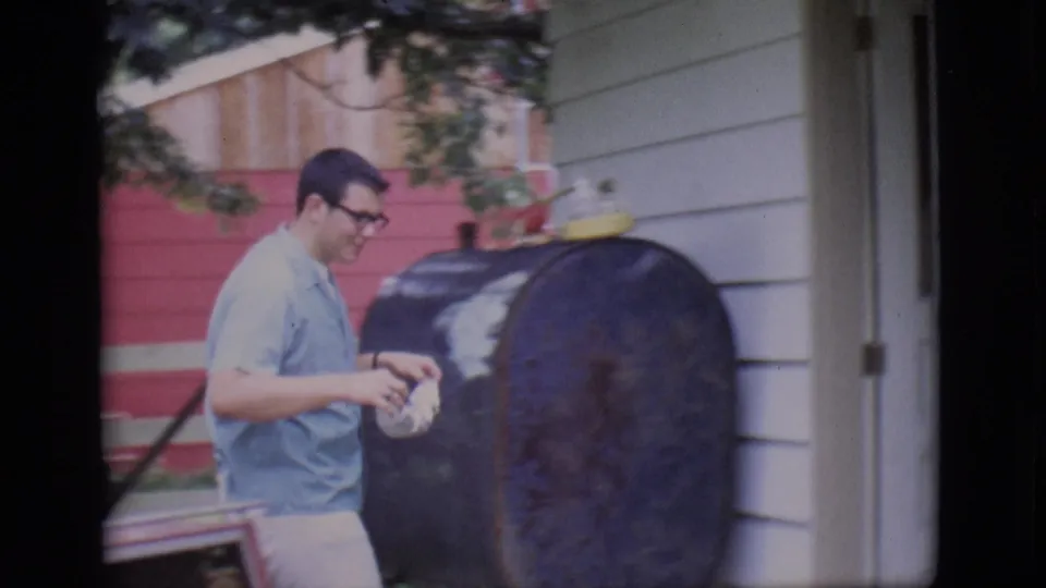 The man holds food beside the black outdoor smoker and red grill in the 1966 Clarkdale backyard footage.
