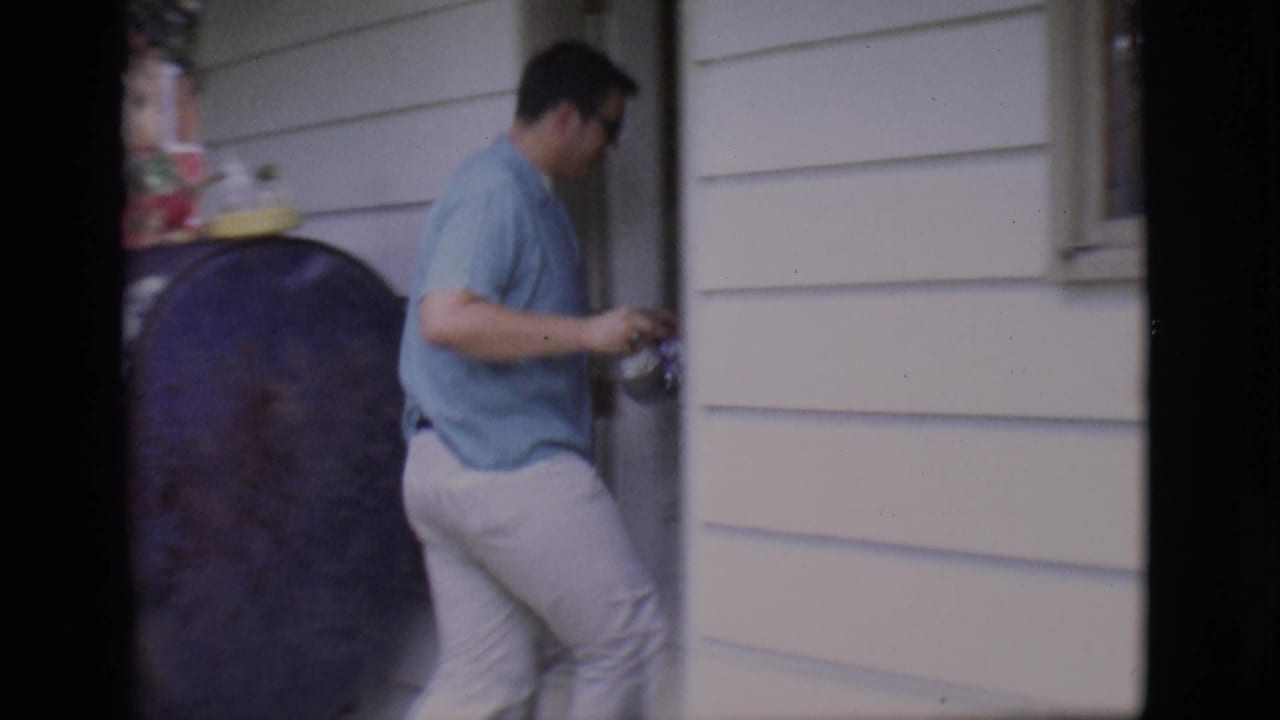 Man in blue shirt stands near a doorway and black tank beside white house siding.