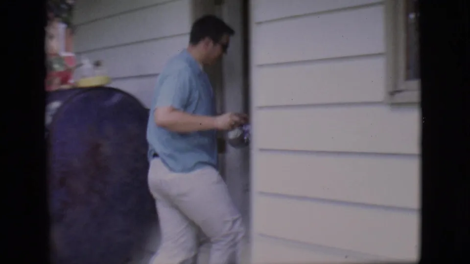 In 1966 Clarkdale home-movie footage, a man in a light blue shirt stands near a doorway and black exterior tank beside white siding.