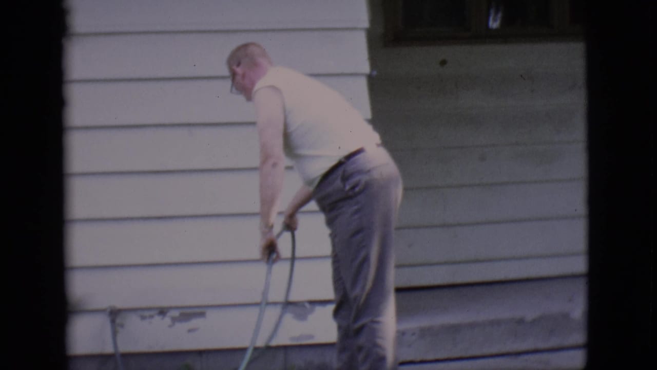 An adult man in a white sleeveless shirt manages a garden hose against the siding of a Clarkdale house in 1966 footage.
